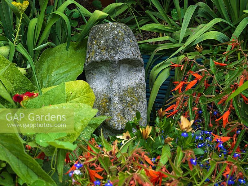 Lichen clad stone head nestles in amongst foliage and flower.