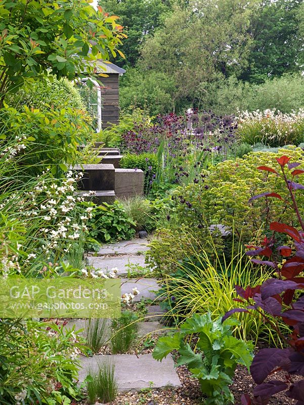 Riverside garden by Andy Sturgeon. Path leads past cotinus, carex, euphorbia, libertia, strawberry, blue fescue to oak cubes by pink allium, erysimum, aquilegia.