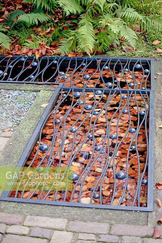 Small square pond filled with floating brown autumn leaves covered with black decorated metal grid in the pavement.