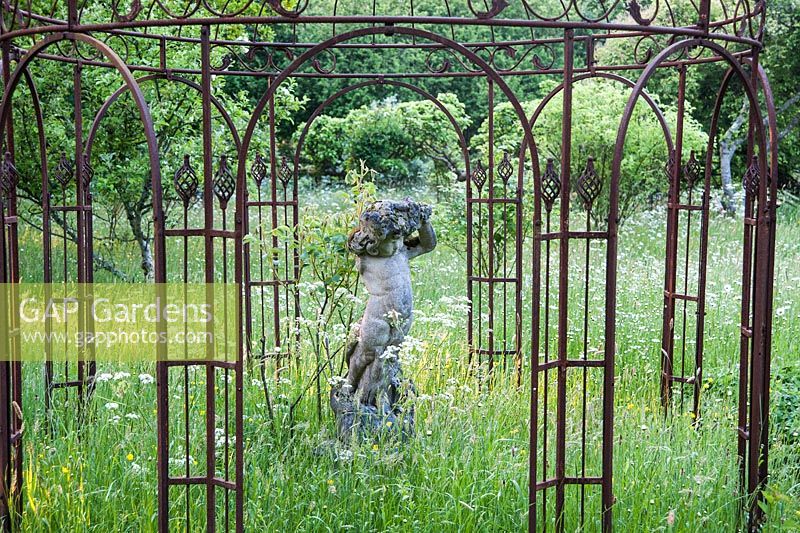 Stone putto framed by an ornate metal arbour  surrounded by meadow full of long grass, buttercups, cow parsley and species roses. King John's Nursery, Etchingham, East Sussex, UK