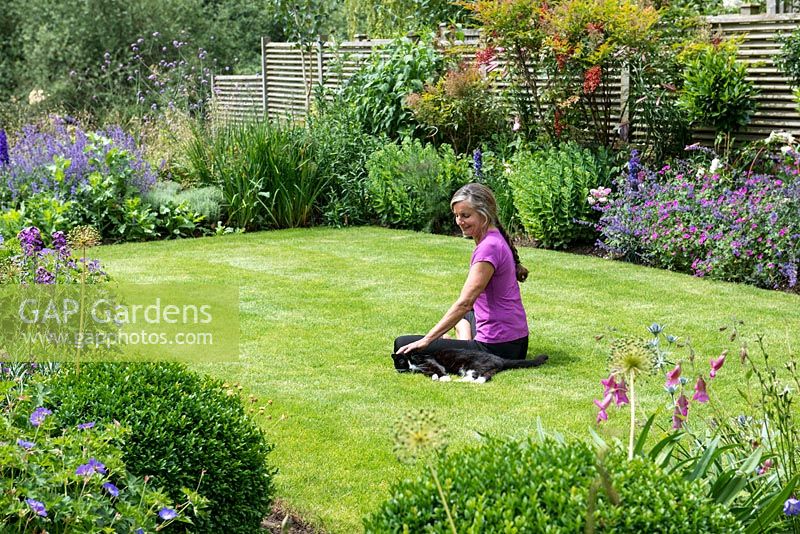 Nicola Stocken relaxes on the 8-week-old lawn in her newly rebuilt riverside garden, four months after it was devastated by February 2014's floods. Lawn enclosed in borders of Geranium Roxanne, box balls, Erysimum Bowles Mauve, delphiniums, Nepeta Walkers' Low, Sedum spectabile, Rosa Harlow Carr, Nandina domestica, Geranium psilostemon.