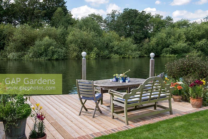 Riverside deck with dining table and chairs, is edged in pots of lillies and herbs, and beds of oriental lilies, daylilies, Verbena bonariensis, annual poppies, agapanthus, dogwood and kniphofia.