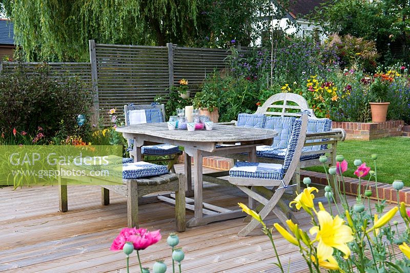 Riverside deck with dining table and chairs, is edged in pots and beds of oriental lilies, daylilies, Verbena bonariensis, annual poppies, agapanthus, dogwood and kniphofia.