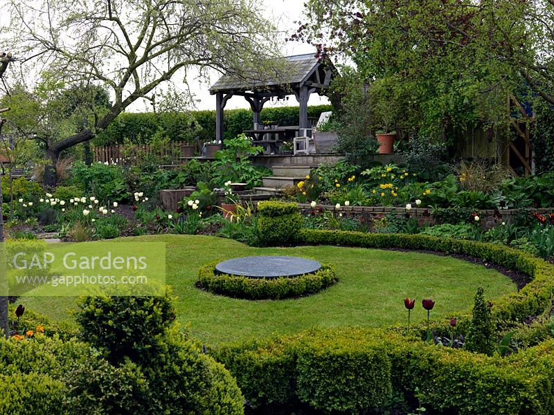 Curving lawn edged in box hedge, at its centre a raised slate circle, a bird table. Terraced beds filled with spring bulbs. Top deck has summerhouse.