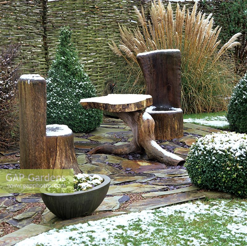 Hidden courtyard with table and seat carved from tree trunk, box topiary and miscanthus, sprinkled with snow.