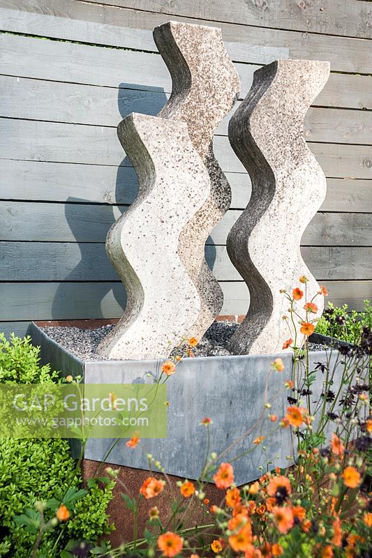 Cast concrete sculpture by Helen Sinclair in the flower garden surrounded by orange and purple geums and aquilegias. 