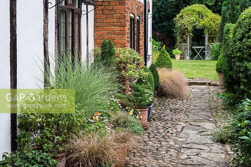 A stone path with terracotta containers planted with ornamental grasses and annuals. In the distance, a clematis covered gazebo with seating area. Southend Farm.