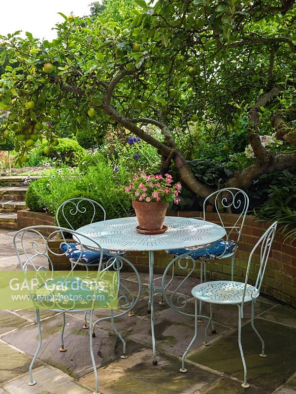 On a sunken terrace enclosed in raised beds, metal bistro table and chairs are shaded by an old apple tree.
