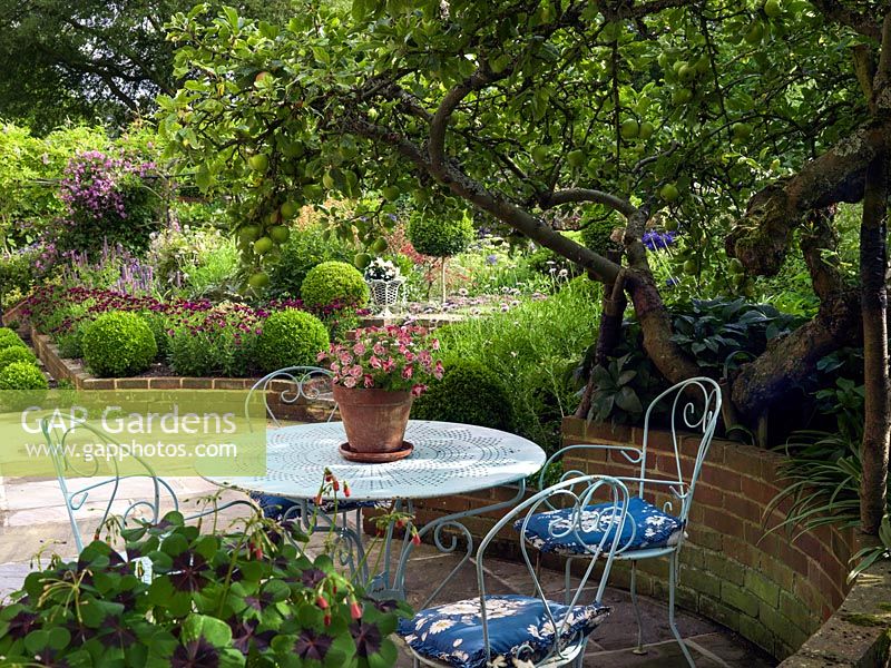 On a sunken terrace enclosed in raised beds, metal bistro table and chairs are shaded by an old apple tree.