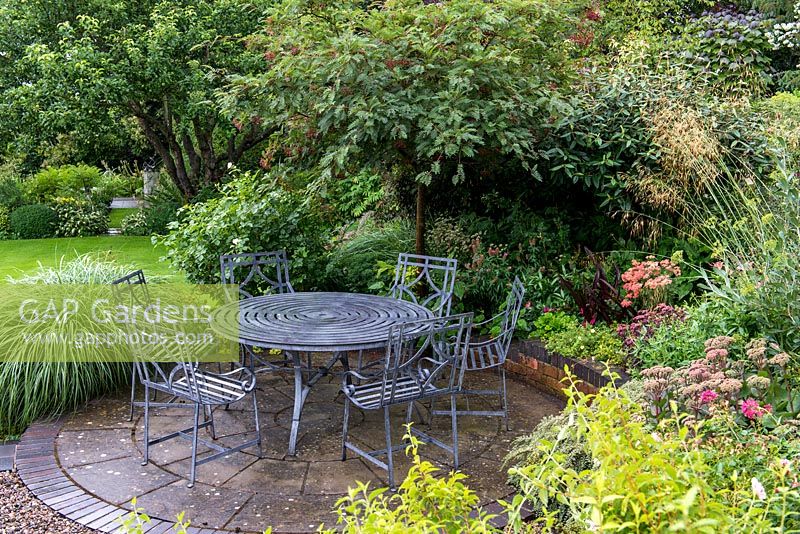 A circular stone patio with table and chairs. Planting in the surrounding raised bed includeds Sedum, Achillea, Stipa gigantea, Viburnum and a Sorbus tree.