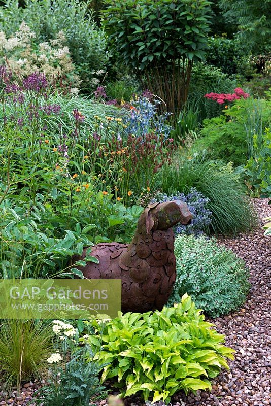 A sculpture of a sheep in metal sits amongst a mixed summer border with plants including with Hosta, Hebe, Geum, Eryngium, Persicaria and Echinacea.