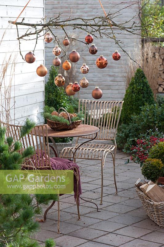 Hung up small furniture of metal, basket with cones and coniferous branches on the table, branch of Malus - apple tree decorated with copper-brown balls over table. Christmas terrace with copper baubles decorating branch