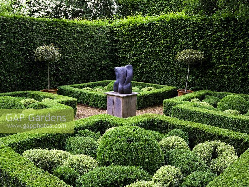 Knot garden, with four box topiary beds grouped around a central sculpture. Enclosed in beech or conifer hedges. Variegated holly standards in pots.