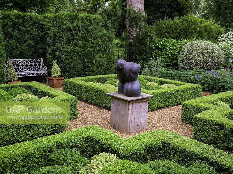 Knot garden, with four box topiary beds grouped around a central sculpture. Enclosed in hedges. White bench in conifer niche. Beyond, community garden.