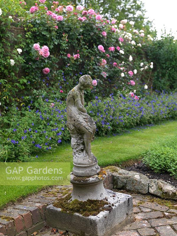 A stone statue stands in a corner of the herb garden. Behind, brick wall bearing climbing Rosa Aloha and Sombreuil - white above hardy geranium.