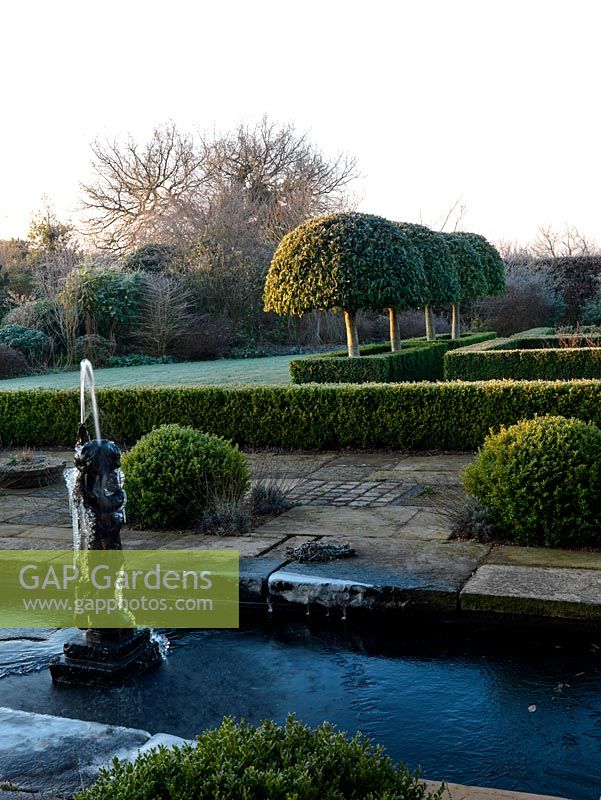 A formal rill fed by a statue fountain. Shaped box balls and hedges with domed tree privet behind.