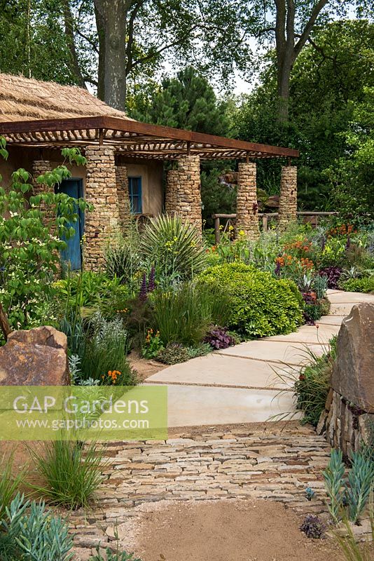 Sentebale - Hope in Vulnerability Garden. View along stone path, past beds planted to reflect the wild landscape of Lesotho and vibrant plant colours, to a Mamohato hut. 