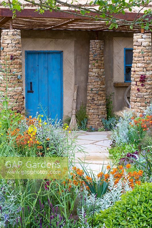 View of the blue door of the grass-roofed hut, with a handmade broom in front. Plants in the foreground include perennial wallflower Erysimum 'Apricot Twist', dyer's woad - Isatis tinctoria, Artemisia ludoviciana 'Valerie Finnis' and Salvia nemorosa 'Caradonna'. Sentebale - Hope in Vulnerability garden. RHS Chelsea Flower Show, 2015