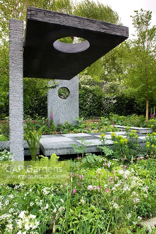 Hard landscaping slate platforms with upright wall panels and one overhead platform with circular holes.  Ferns, aquilegia, euphorbia. Shade planting mostly green. RHS Chelsea Flower Show 2015