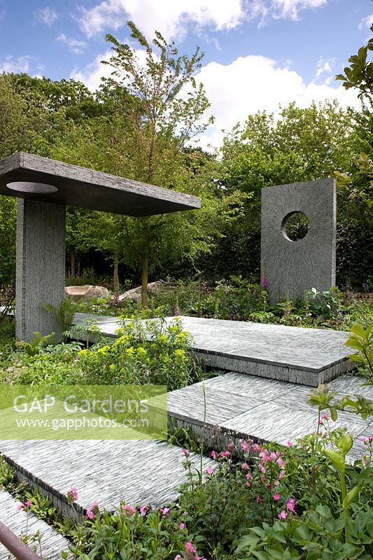 Slate platforms with upright wall panels and one overhead platform with circular holes.  Ferns, aquilegia, euphorbia. Shade planting mostly green. The Brewin Dolphin Garden. RHS Chelsea Flower Show, 2015