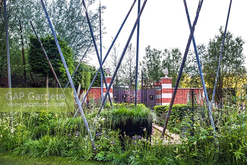 view of steel poles, walls of red brick and Bath stone with ornamental iron gate surrounded by mixed planting. The Living Legacy Garden, RHS Chelsea Flower Show 2015