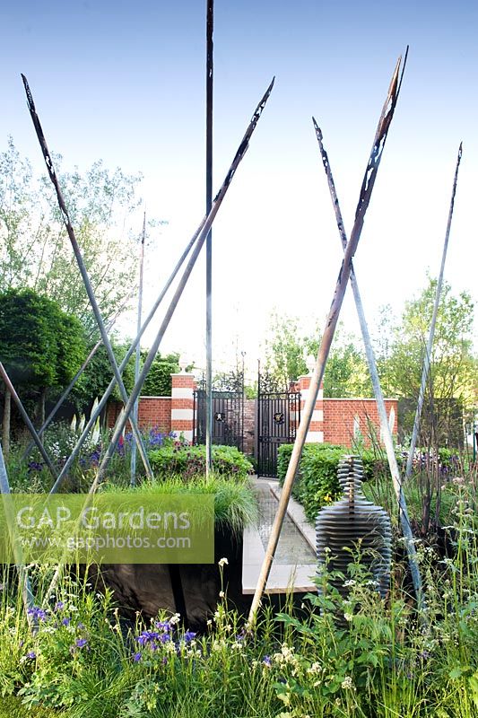 Formal gates from Wellington College behind planting of Beech hedge, Eremurus and grasses with pole sculpture and figure representing the Battle of Waterloo. Water rill in centre axis. The Living Legacy Garden by Darwin Property Management Investment. RHS Chelsea Flower Show, 2015