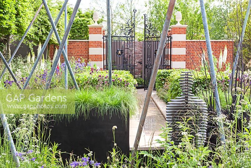 View through the garden from the sculpture of a figure through the metal poles and the rill to the gated entrance to the courtyard with the initials 'A' and 'W' on the gates The Living Legacy Garden by Darwin Property Management Investment. RHS Chelsea Flower Show, 2015