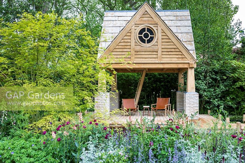 View of two storey oak framed summerhouse with ladder and fabric chairs, wooden boardwalk next to swimming pond. Planting in the foreground features an Acer - maple, M and G garden. RHS Chelsea Flower Show, 2015.