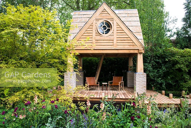 building, wooden deck patio with two chairs and bridge over pond surrounded by flowerbeds with Rosa 'Nuits de Young', Rosa 'Chianti', Salvia nemorosa 'Caradonna' and Verbascum 'Cotswold beauty' and Acer - The M and G Garden. RHS Chelsea Flower Show 2015