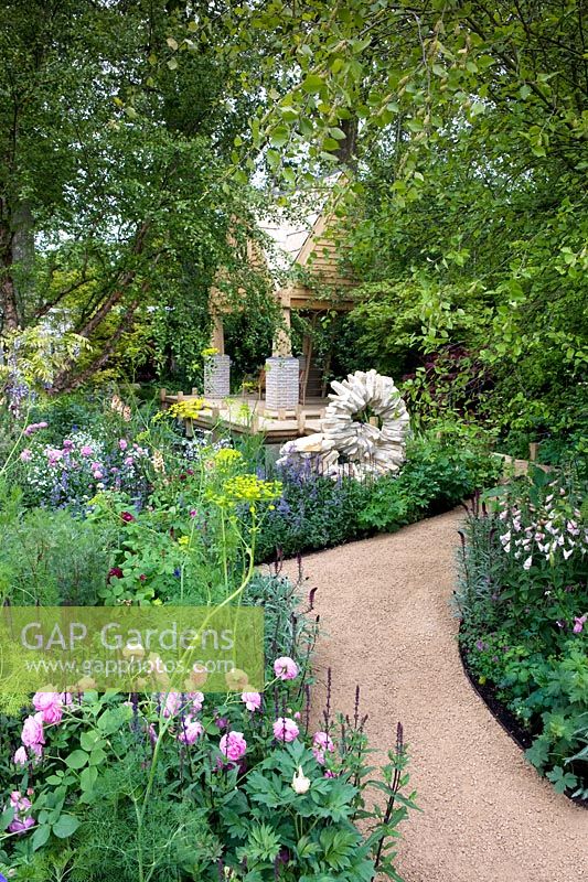 The M and G Garden - The Retreat. View down winding path to oak-framed garden building with stone sculpture. Cottage-style planting with roses and foxgloves. Rosa Louise Odier