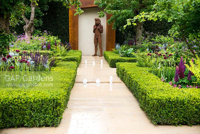 The Morgan Stanley Healthy Cities Garden. View of sandstone path with water feature leading to steel sculpture man with a child surrounded by symmetric hedges - Buxus sempervirens, fern - Dryopteris affinis, Salvia nemorosa 'Caradonna',  Camassia cusickii and Lupinus 'Masterpiece'.