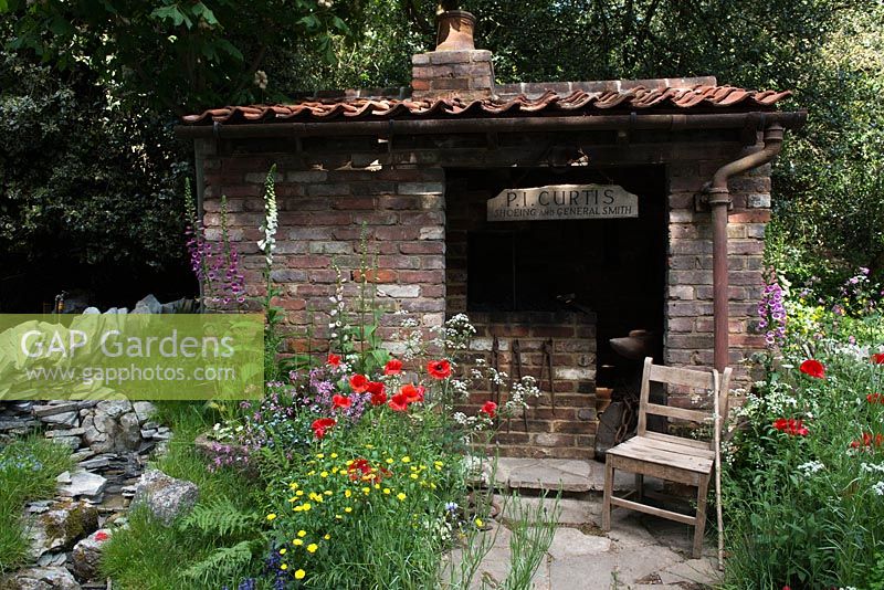 View of natural rock waterfall and vintage brick shed surrounded by wild flowers Digitalis purpurea, Papaver rhoeas, Anthriscus sylvestris, Ranunculus acris. The Old Forge Artisan Garden for Motor Neurone Disease Association - RHS Chelsea Flower Show 2015. 

