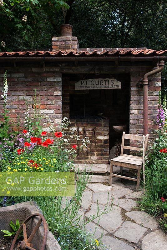The Old Forge Artisan Garden for Motor Neurone Disease Association - RHS Chelsea Flower Show 2015. View of natural rock pathways and a brick shed and vintage chair surrounded by wildflowers