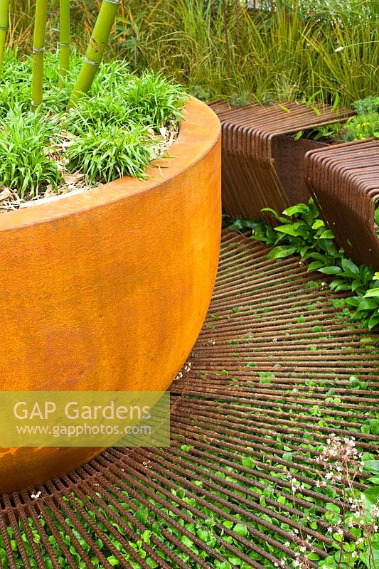 A rusted corten steel container planted with Phyllostachys and grasses positioned on a metal grid with adjacent seats. Dark Matter Garden for the National Schools' Observatory.  RHS Chelsea Flower Show, 2015. 