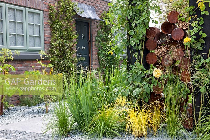 The Great Chelsea Garden Challenge. Border with ornamental grasses, yellow flowering roses, rusted steel cans with bee hotels, gravel and stone paving.