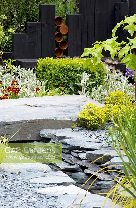 View across hard landscaping Reclaimed stone paving and water channel with plants euphorbia, box, stachys and angelica.  The Great Chelsea Garden Challenge Garden.  RHS Chelsea Flower Show, 2015. 