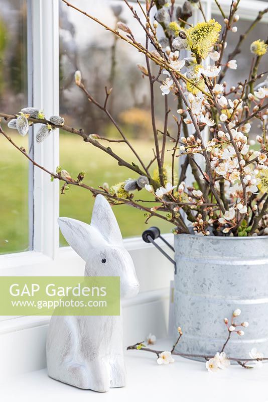 Floral display containing blossoming spring foliage in a galvanised bucket, a bunny and a view to the garden beyond