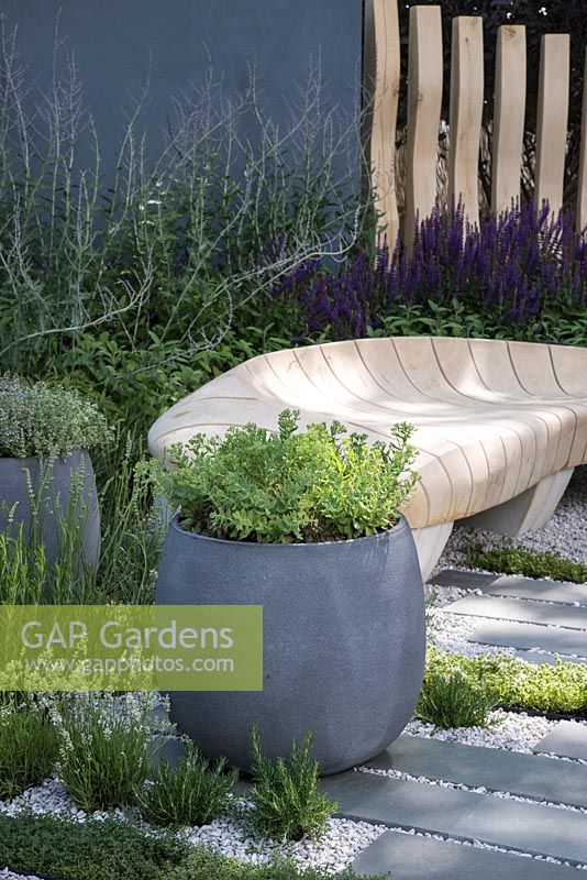 Container planted with sedum, curved oak bench, paving with Thymus argenteus, Rosmarinus officinalis and Lavandula, border planting of Salvia nemorosa and Perovskia - Living Landscapes: Healing Urban Garden, RHS Hampton Court Palace Flower Show 2015