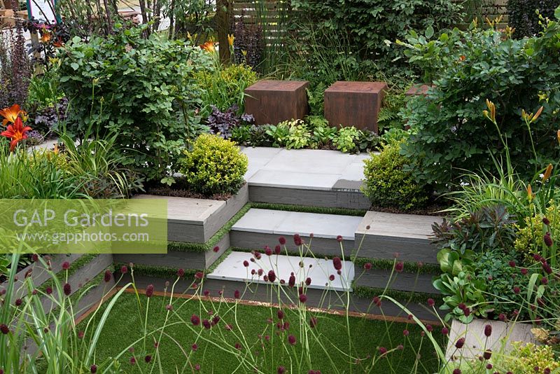 Metal cubed seating with steps down to sunken garden and artificial lawn, Sanguisorba officinalis 'Morning Select' in foreground - Foundation for Growth Garden, RHS Hampton Court Palace Flower Show 2015