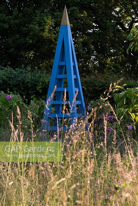 Decorative blue wooden obelisk in the rose garden, grasses in evening light. Heveningham, June