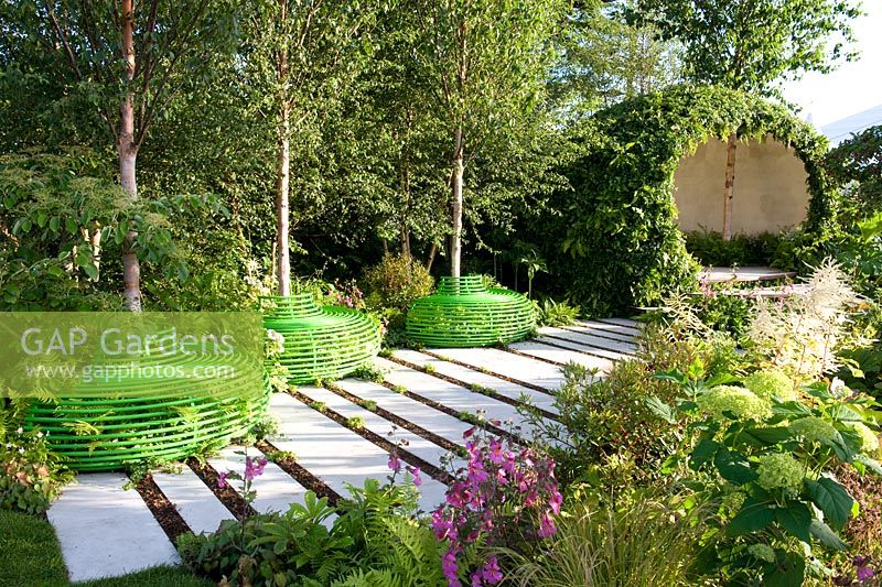  White stone paving through shady woodland-edge garden with pod structure seating area. Three birch trees with green-painted seats. The Macmillan Legacy Garden Sponsor Macmillan Cancer Support, RHS Hampton Court Palace Flower Show 2015