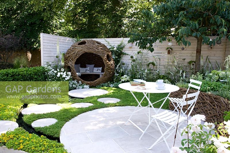 Wildlife-friendly small urban garden with chamomile lawn, circular paving, a woven willow bird-hide seat and table and chairs. White fence.  Living Landscapes. City Twitchers.  Silver Gilt Medal Summer Garden 