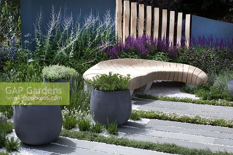 Sculptured bench, striped paving interspersed with herb stripes, and pots of herbs in Living Landscapes: Healing Urban Garden at RHS Hampton Court Palace Flower Show 2015
