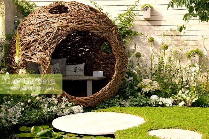 Spherical woven willow bird hide seating area in wildlife-rich garden with Chamomile lawn and circular stepping stones and white and green flower beds with Eryngium giganteum, Digitalis purpurea albiflora, Hydrangea macrophylla, Cimicifuga racemosa, Rosa, Hosta Fire and Ice, Acanthus mollis, Campanula persicifolia Alba in Living Landscapes: City Twitchers garden at RHS Hampton Court Palace Flower Show 2015.