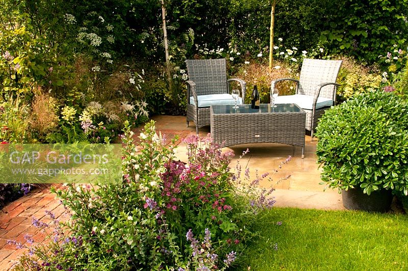 Brick path leading to secluded rectangular seating area with ratten chairs and table and surrounded by flowerbeds with mixed planting in A Garden by Association at RHS Hampton Court Flower Show 2015. A garden designed and built by members of The Association of Professional Landscapers 