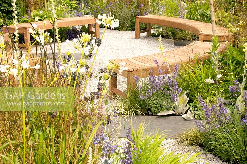 Circular seating area on gravel with curving wooden benches and surrounded by yellow white and blue flower beds with Agapanthus africanus Albus Cerinthe major Purpurascens Eryngium x zabelii Nepata racemosa Walker's Low Cosmos Verbascum bombyciferum and grasses in the Unique: The Rare Chromosome Disorder Garden at RHS Hampton Court Flower Show 2015 