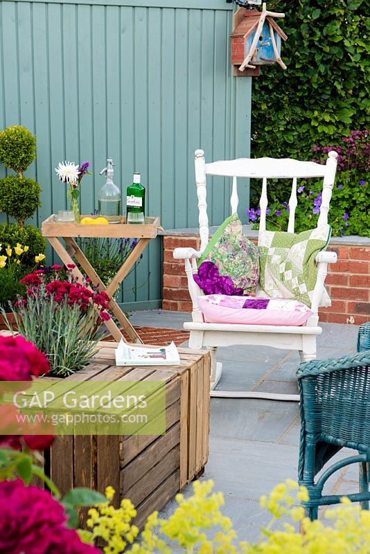 Just Retirement: A Garden For Every Retiree, view of seating area with white rocking chair, table made from wooden crates against blue wall and brick raised flower bed surrounded by red Dianthus, Roses. Designer: Tracy Foster Sponsor:  Just Retirement Ltd