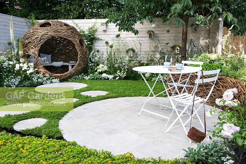 City Twitchers Garden -  view of woven willow bird hide and seating area with metal white furnitures and stone - concrete circular slabs as a path over a pond surrounded by Chamaemelum nobile - chamomile lawn and Sedum carpet. 