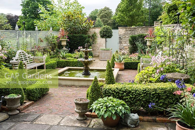 Formal courtyard garden behind Georgian terraced house with central water feature, box edged beds infilled with Japanese anemones, echinops and roses, box cones, stone urns, a standard bay and containers planted with succulents and agapanthus.