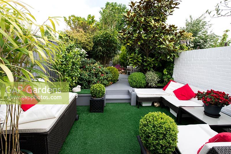 3.7m x 25m contemporary garden. 3 seating areas are linked by stepped plank path. Borders of evergreens include box balls, magnolia, bamboo, and rhododendron. Artificial grass lawn.